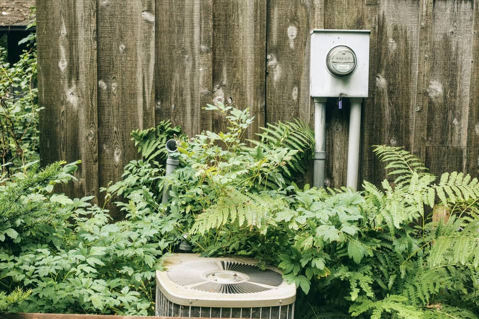 An outdoor air conditioning condenser among ferns, below an electric meter on a wooden wall